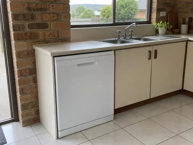 Built-in dishwasher under a kitchen bench beside a sink, with tiled floor and brick wall in Mount Hutton